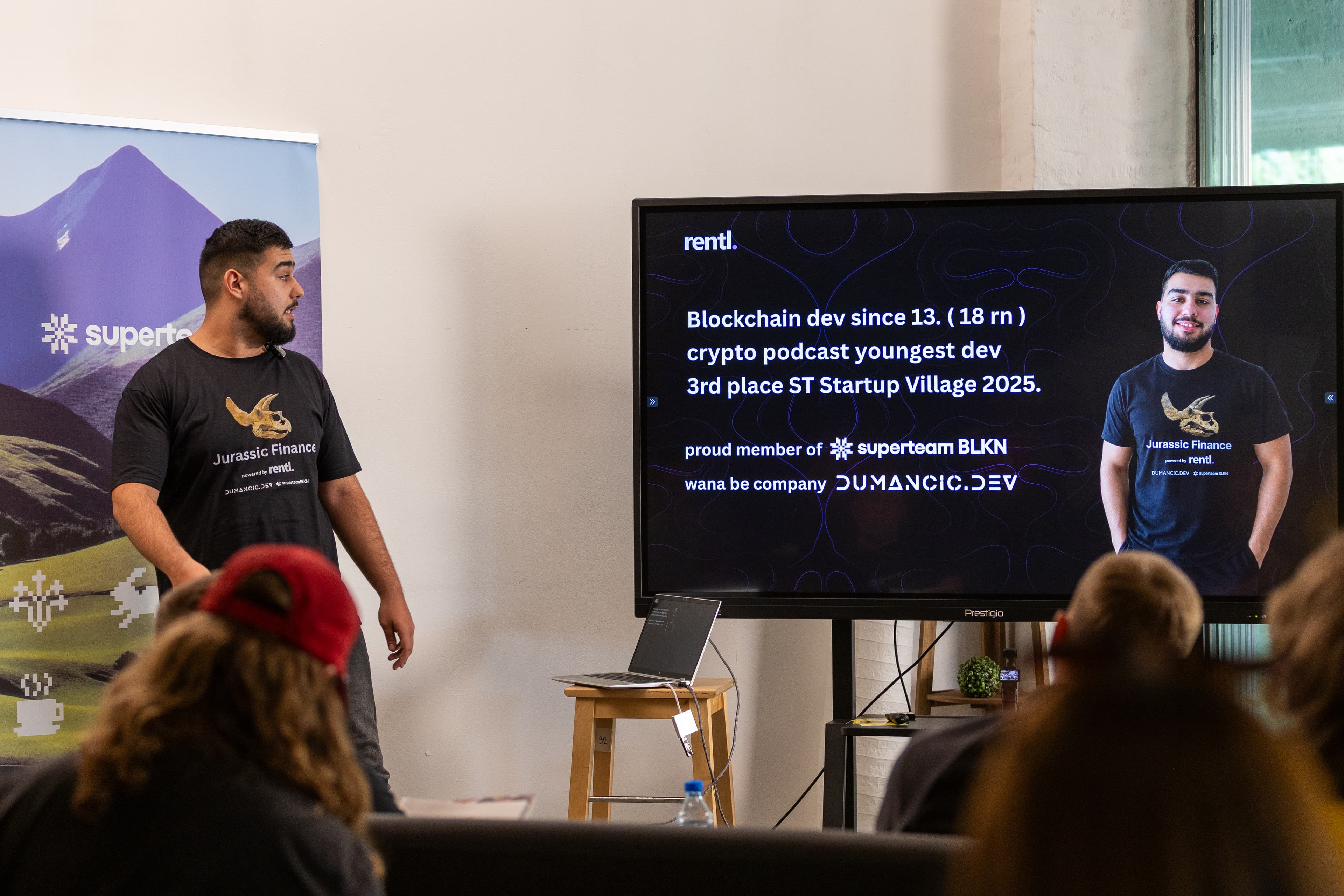 Toni on stage at Superteam Balkan Demo Day in Belgrade, wearing the auctioned Jurassic Finance t-shirt in front of the audience, with the Rentl pitch slide on the screen beside him.